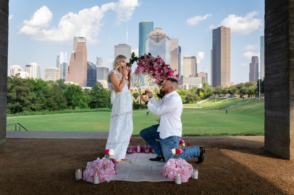 Surprise proposal in Houston park with man proposing to girlfriend under romantic floral arrangement and skyline backdrop