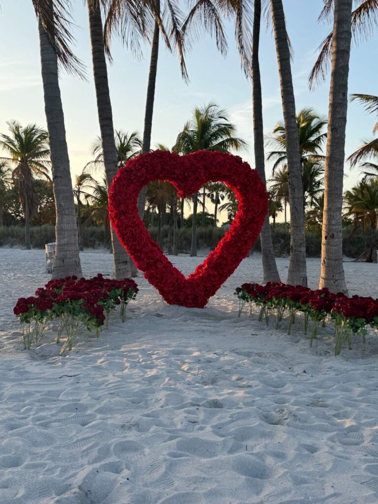 Miami Beach proposal setup with large red rose heart arch on the sand at sunset surrounded by palm trees
