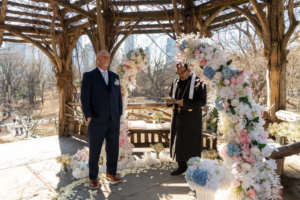 Groom and officiant during a Central Park winter elopement ceremony beside a lush pink and blue floral arch