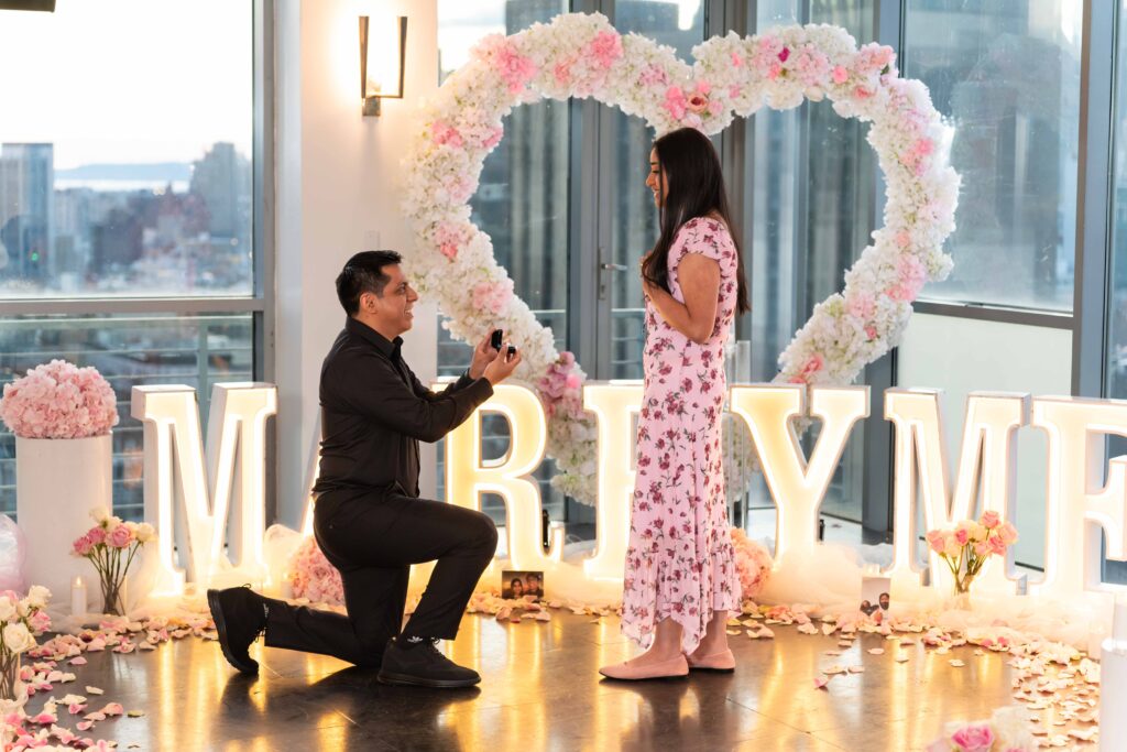 Man proposing indoors in NYC with a floral heart arch and illuminated Marry Me letters